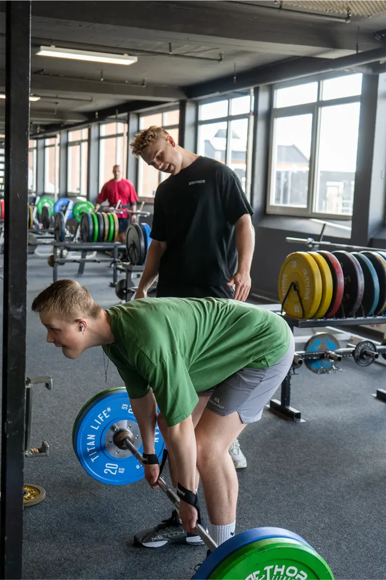 Træner vejleder klient under dødløft med vægtstang i fitnesscenter.